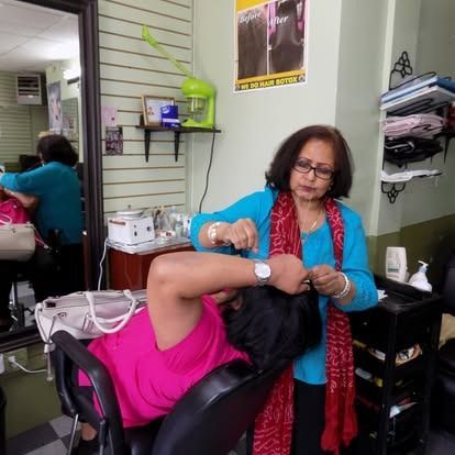 Woman styling another woman's hair in a salon. The stylist wears blue; the client, pink.