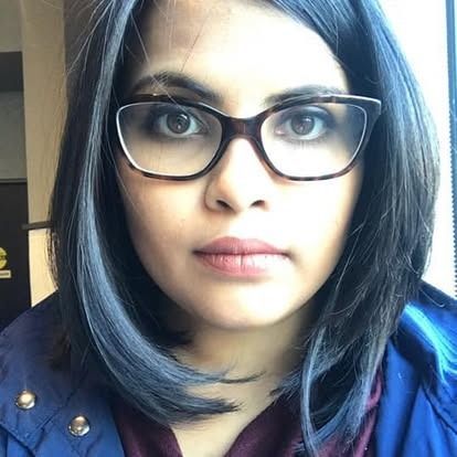 Woman with dark hair and glasses, looking directly at the camera. Wearing blue jacket.