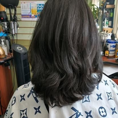 Person with dark, layered hair at a salon, wearing a patterned cape.