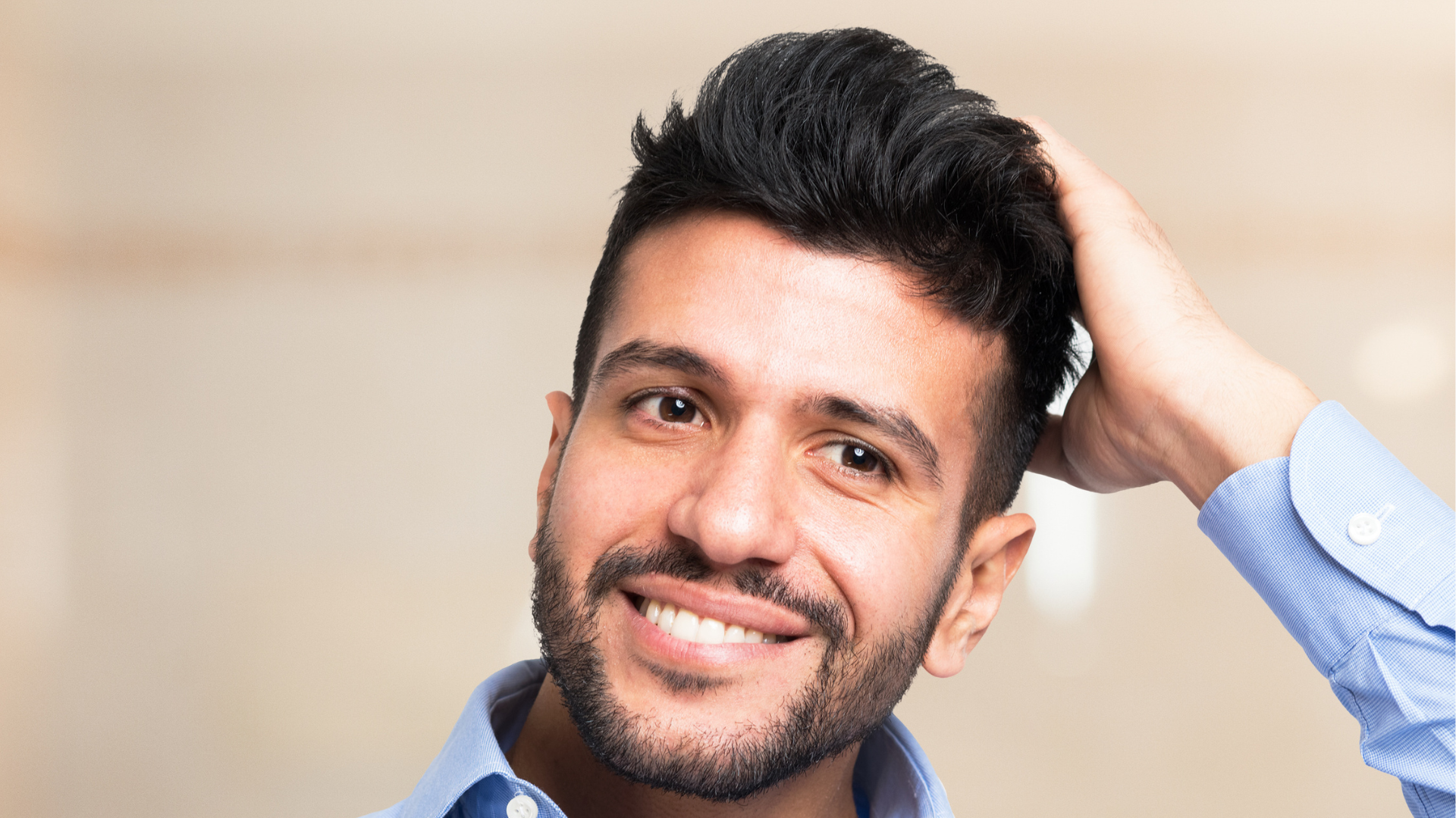 Smiling man with dark hair, touching his hair, wearing a blue shirt, blurred background.