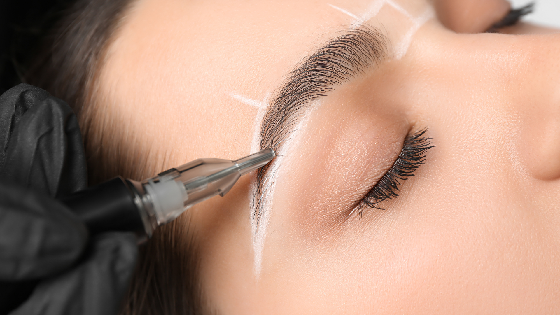 Person receiving eyebrow microblading, close-up; technician's gloved hand holding a needle.