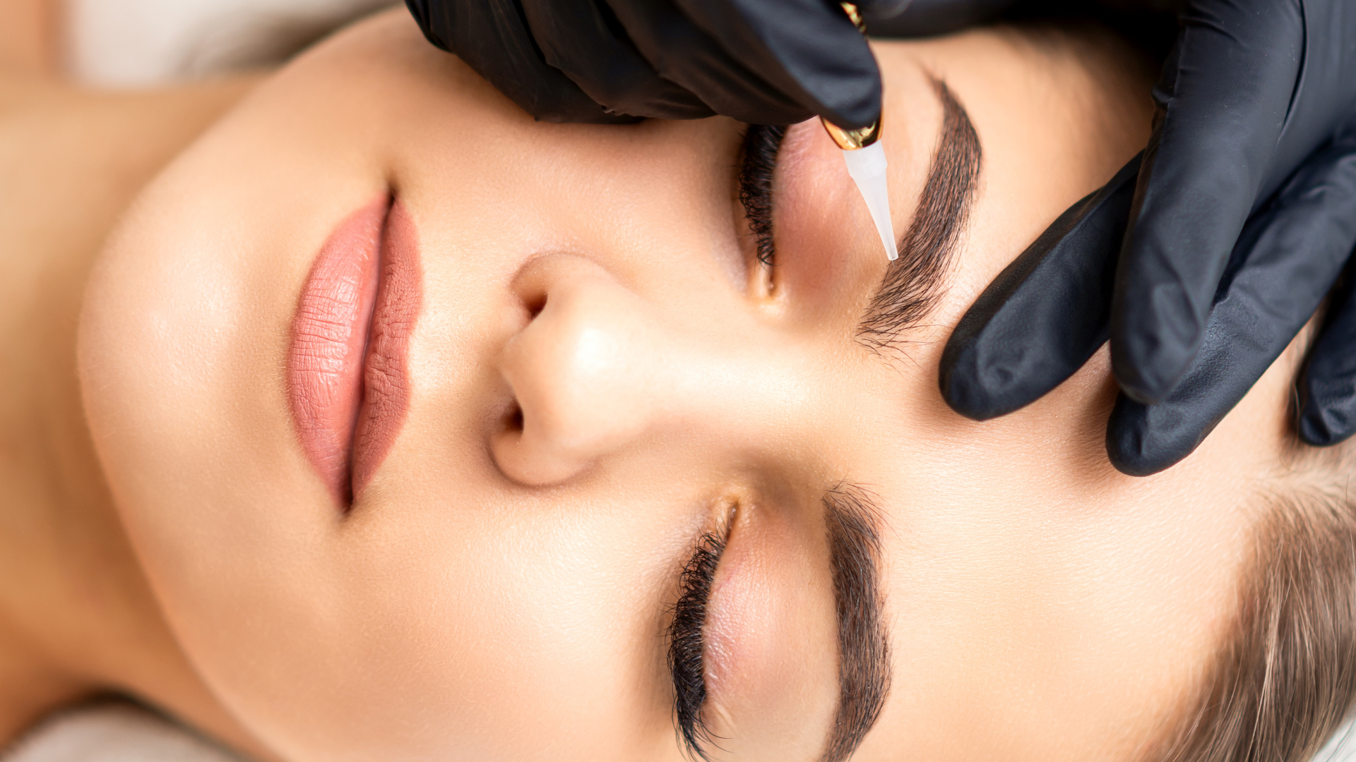Person receiving microblading on eyebrows, hands in black gloves.