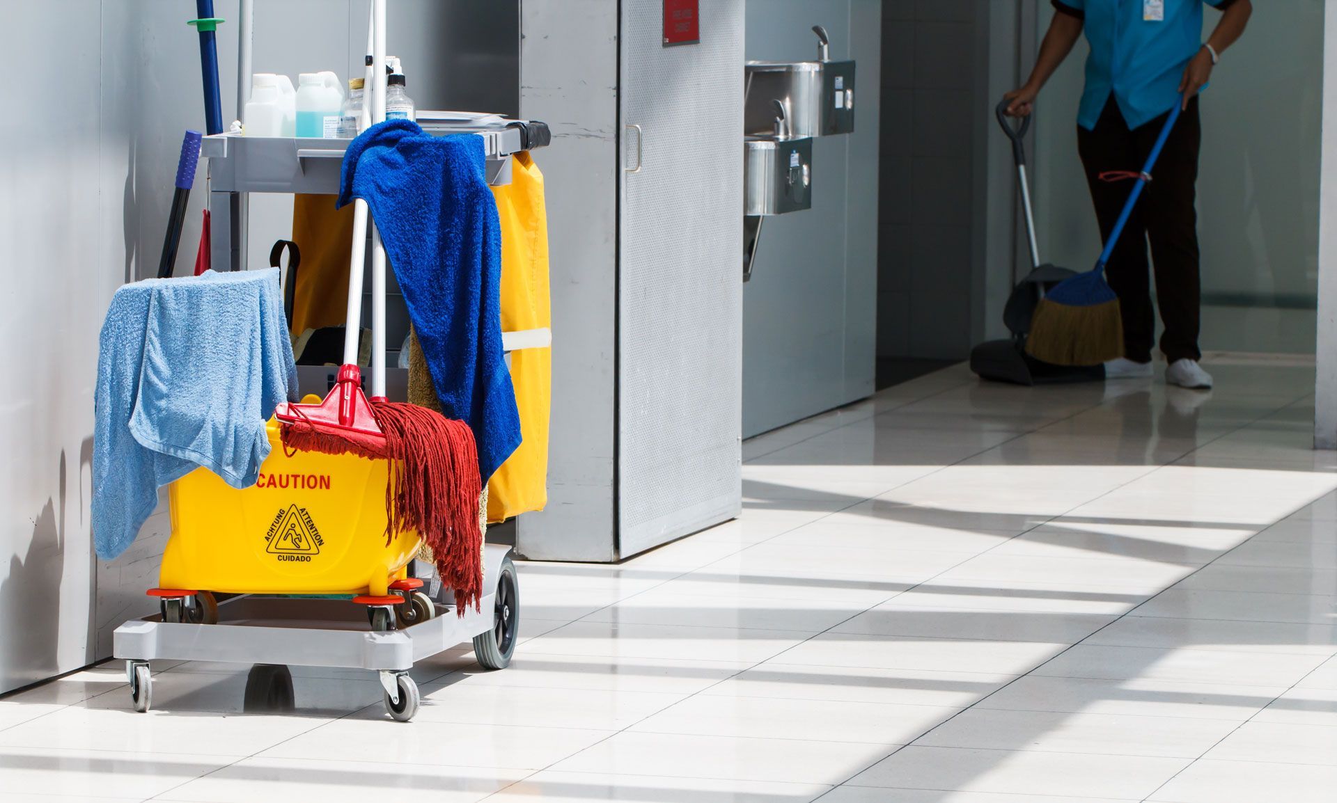A man is cleaning a hallway with a mop and a cart of cleaning supplies.
