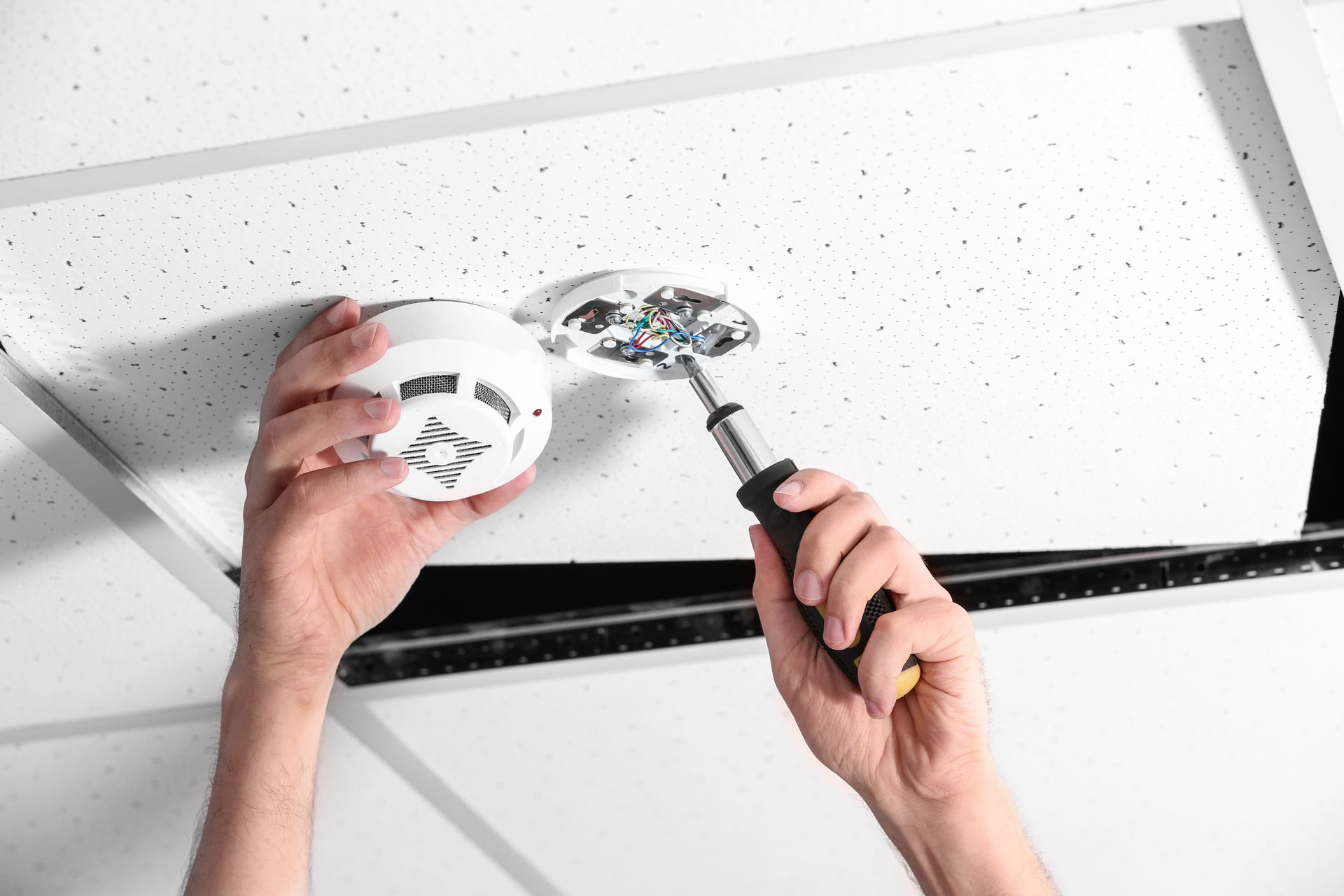 Hands installing a smoke detector on a ceiling tile with a screwdriver