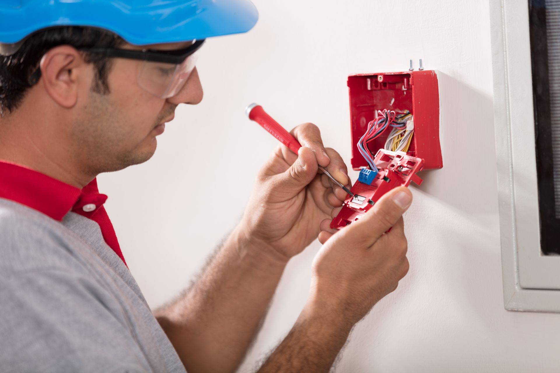 Un ouvrier installe un boîtier de prise électrique rouge sur un mur, portant un casque de chantier bleu et des lunettes de sécurité.