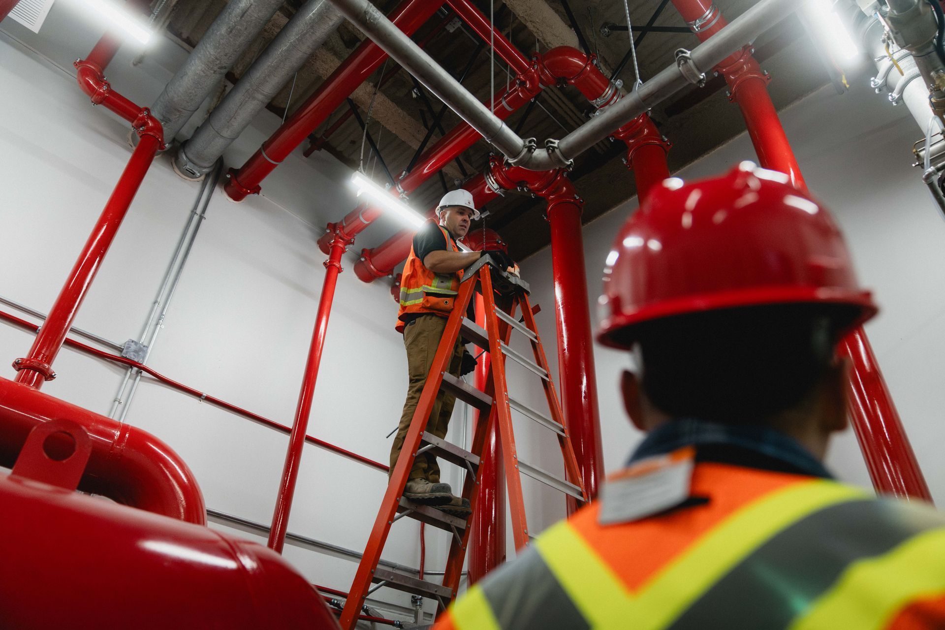 Two workers in hard hats inspecting red fire pipes in an industrial room, one on a ladder