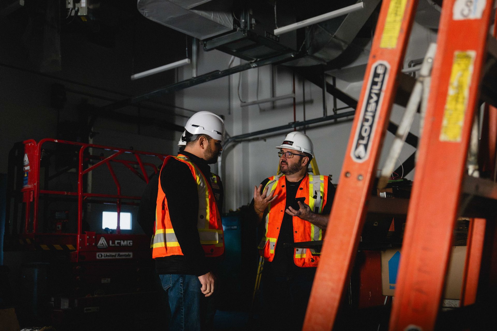 Two construction workers in hard hats and safety vests talk beside orange ladders in a dim industrial site