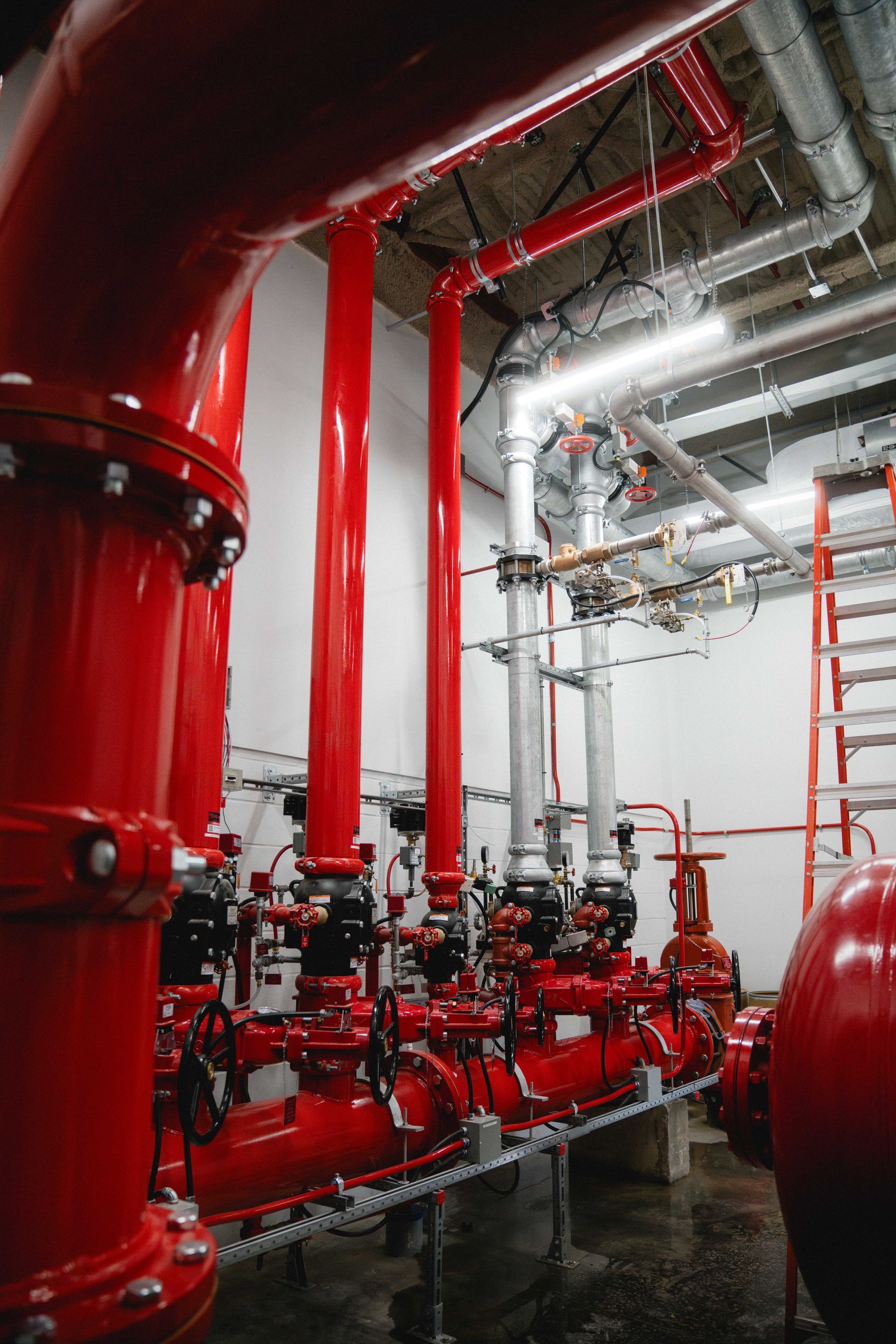 Red industrial pipes and valves in a mechanical room with a red ladder and ceiling ductwork