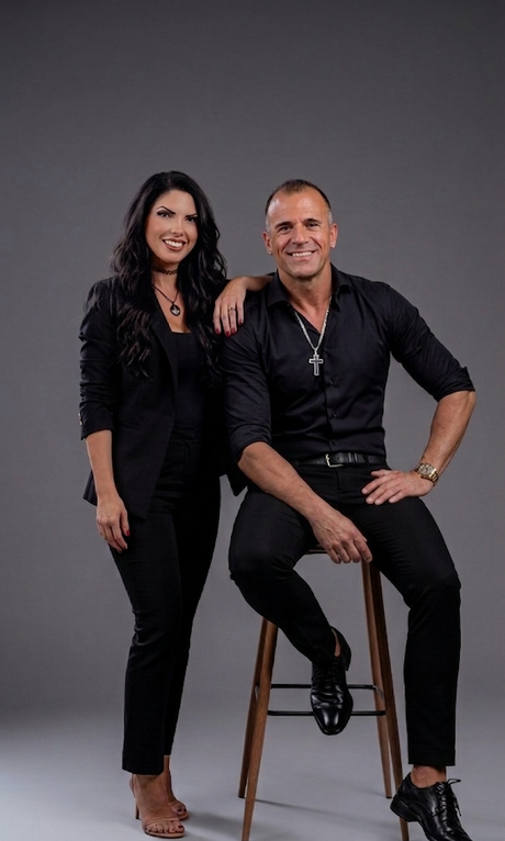 Two people posing against a gray backdrop, one seated on a stool and one standing beside them.