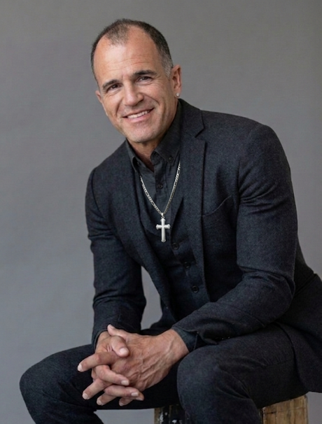 Smiling man in a dark suit with a cross necklace, seated against a gray backdrop.