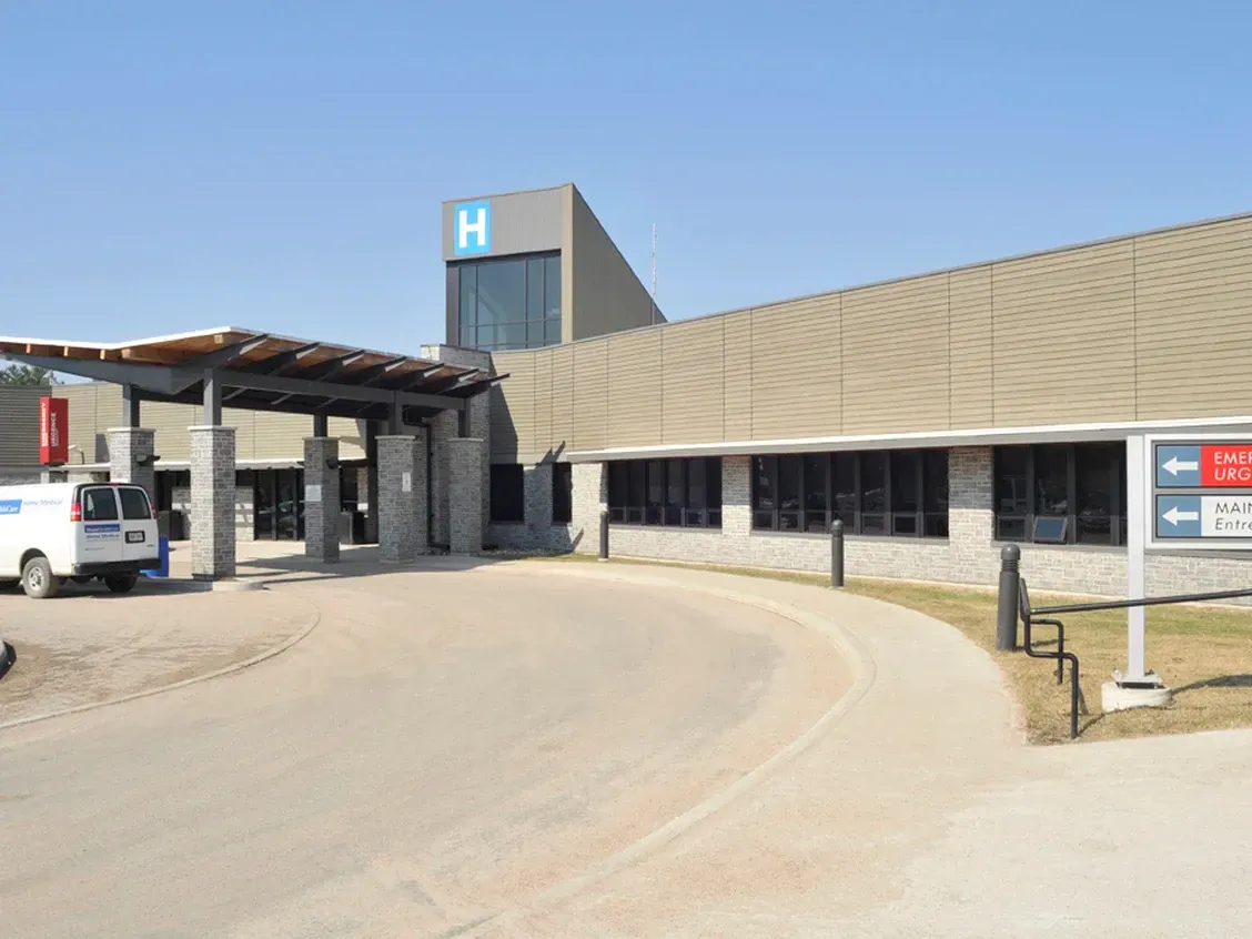 A modern, single-story hospital building with a grey stone exterior, a covered entrance, and a white van parked nearby.