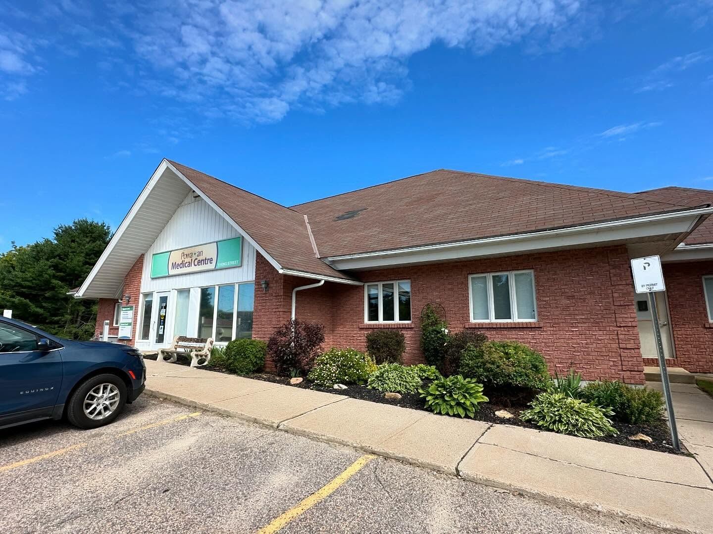A one-story red brick building with a brown roof under a blue sky, featuring a storefront entrance and a parking lot.