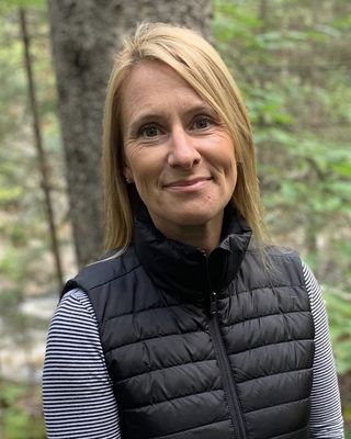 A smiling blonde woman in a black puffer vest and striped long-sleeve shirt, standing outdoors in front of a forest.