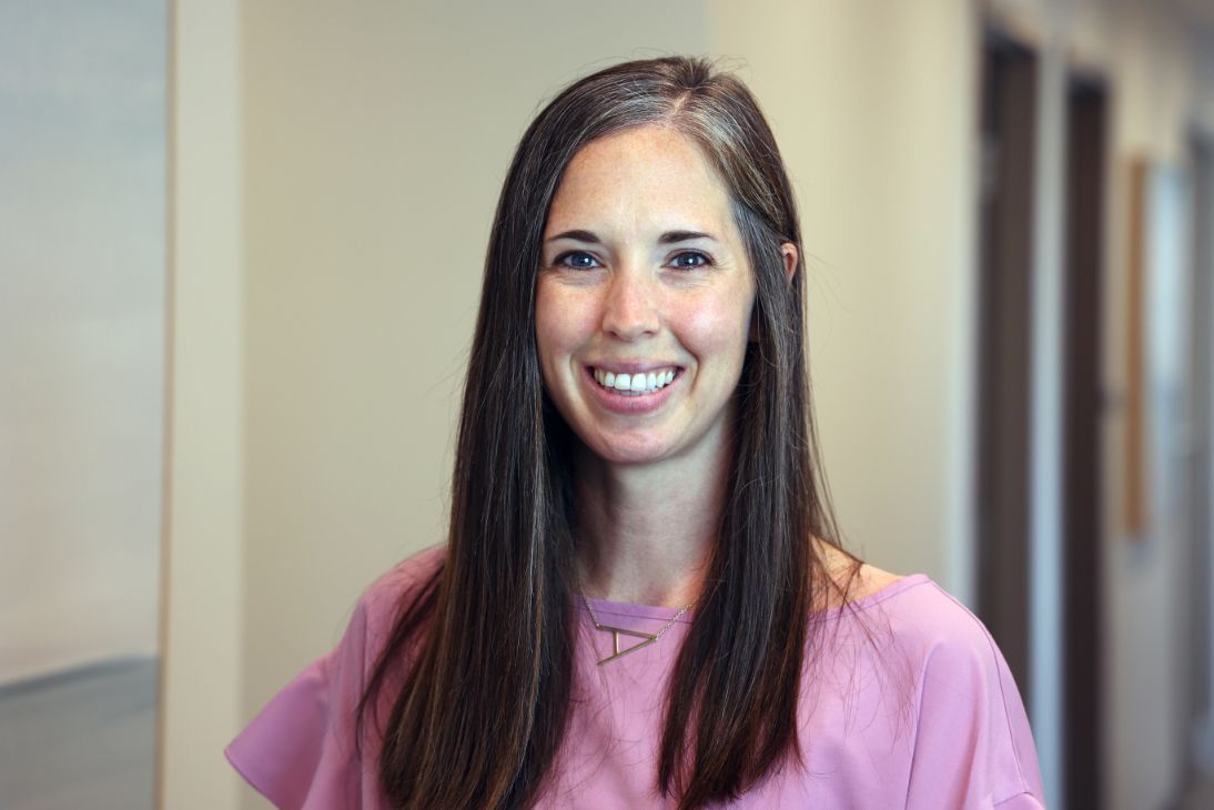 A smiling person with long dark hair, wearing a pink top, stands in a brightly lit indoor hallway.