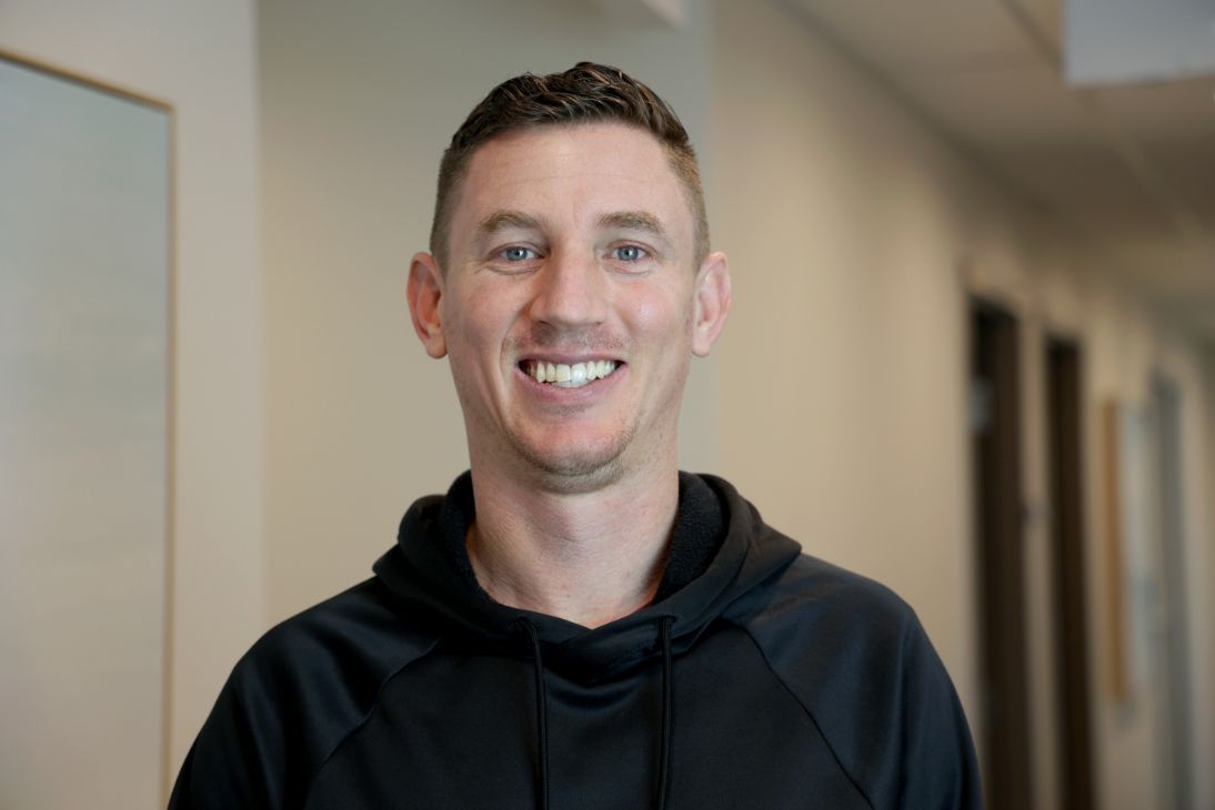 A person wearing a black hooded sweatshirt smiles warmly while standing in a brightly lit, professional hallway.