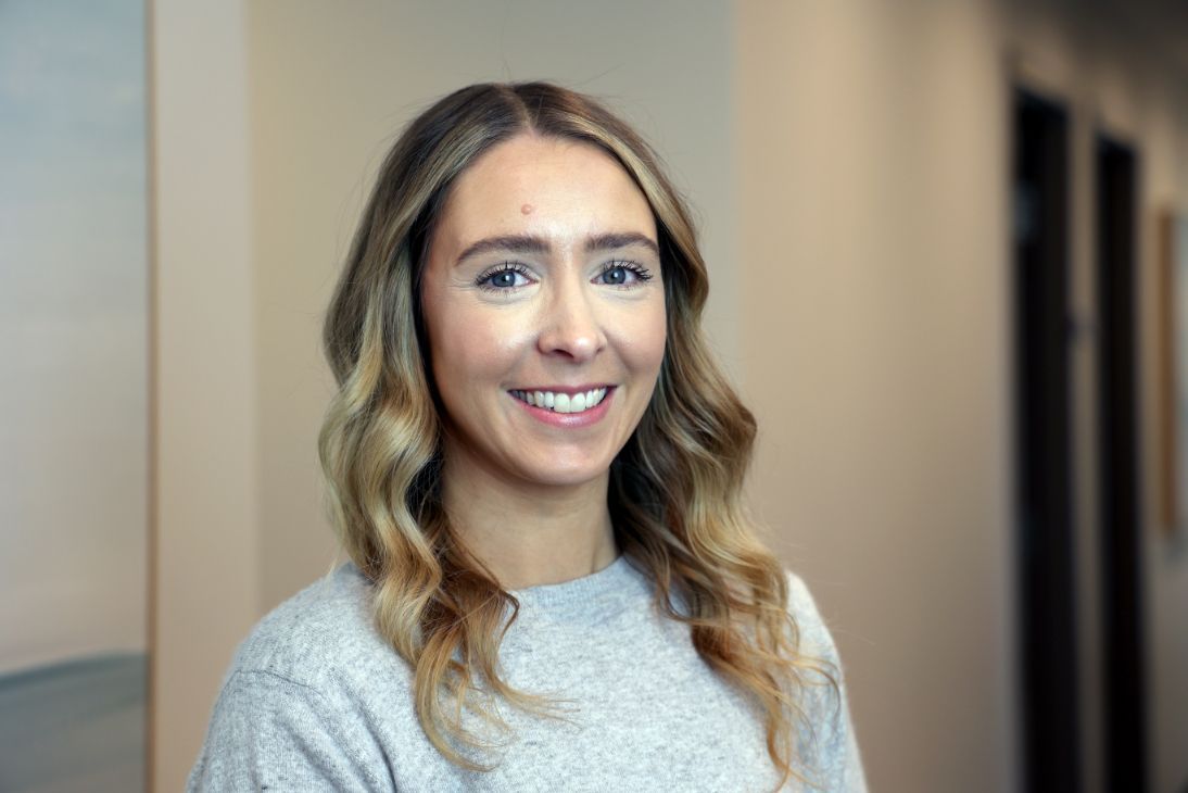 A person with long, wavy light brown hair smiling at the camera, wearing a light gray sweater in an indoor setting.