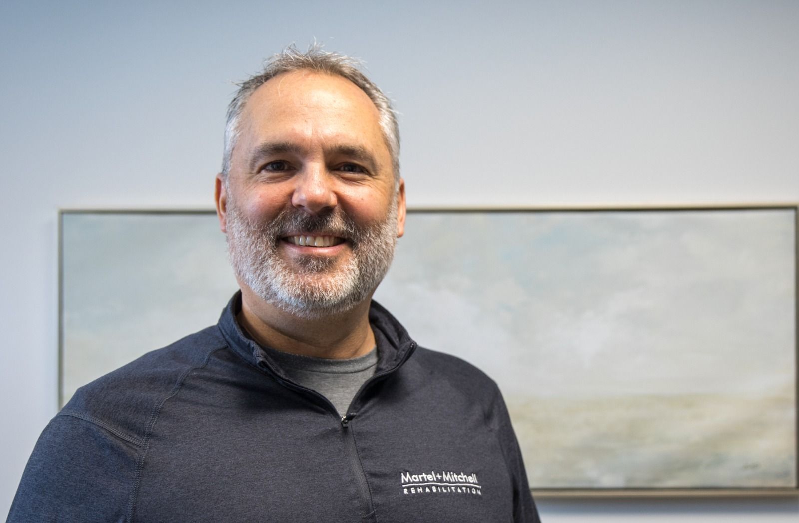 A man with short grey hair and a beard, smiling warmly while wearing a black athletic jacket.