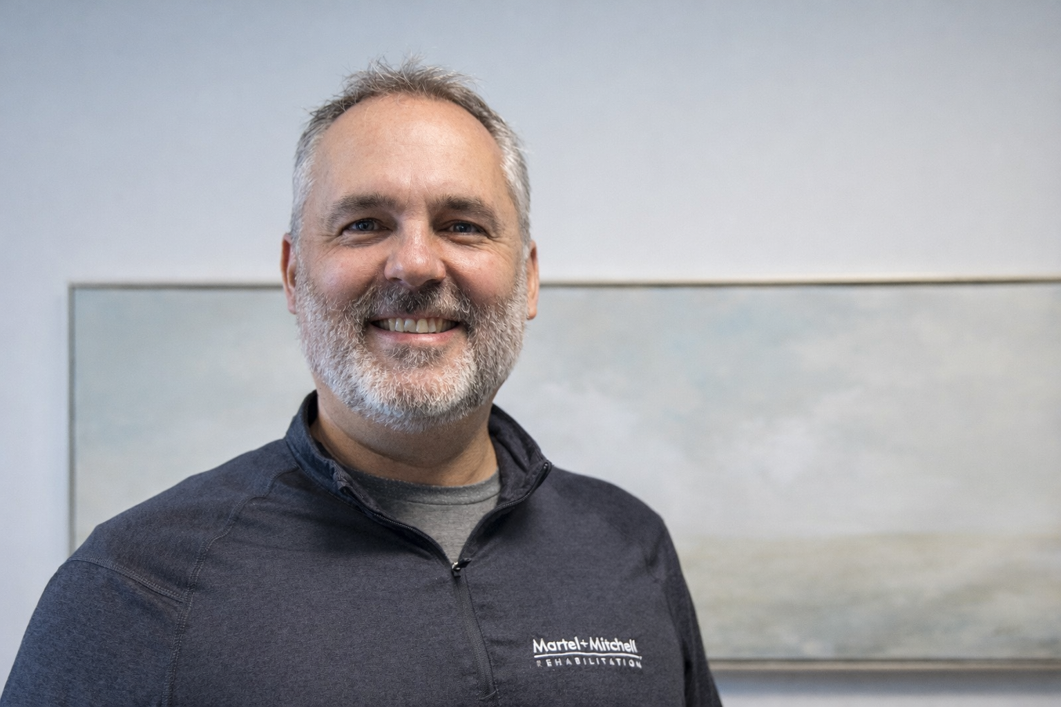 A man with short grey hair and a beard, smiling warmly while wearing a black athletic jacket.