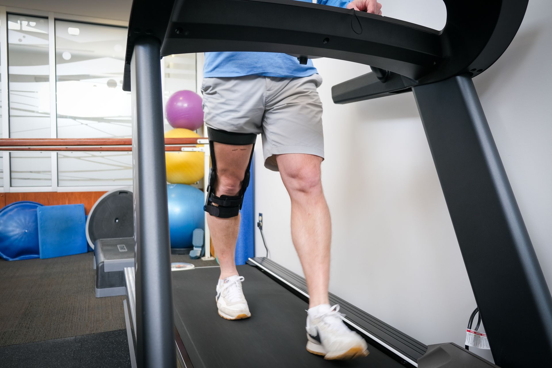 A person wearing a leg brace walks on a treadmill in a physical therapy gym.