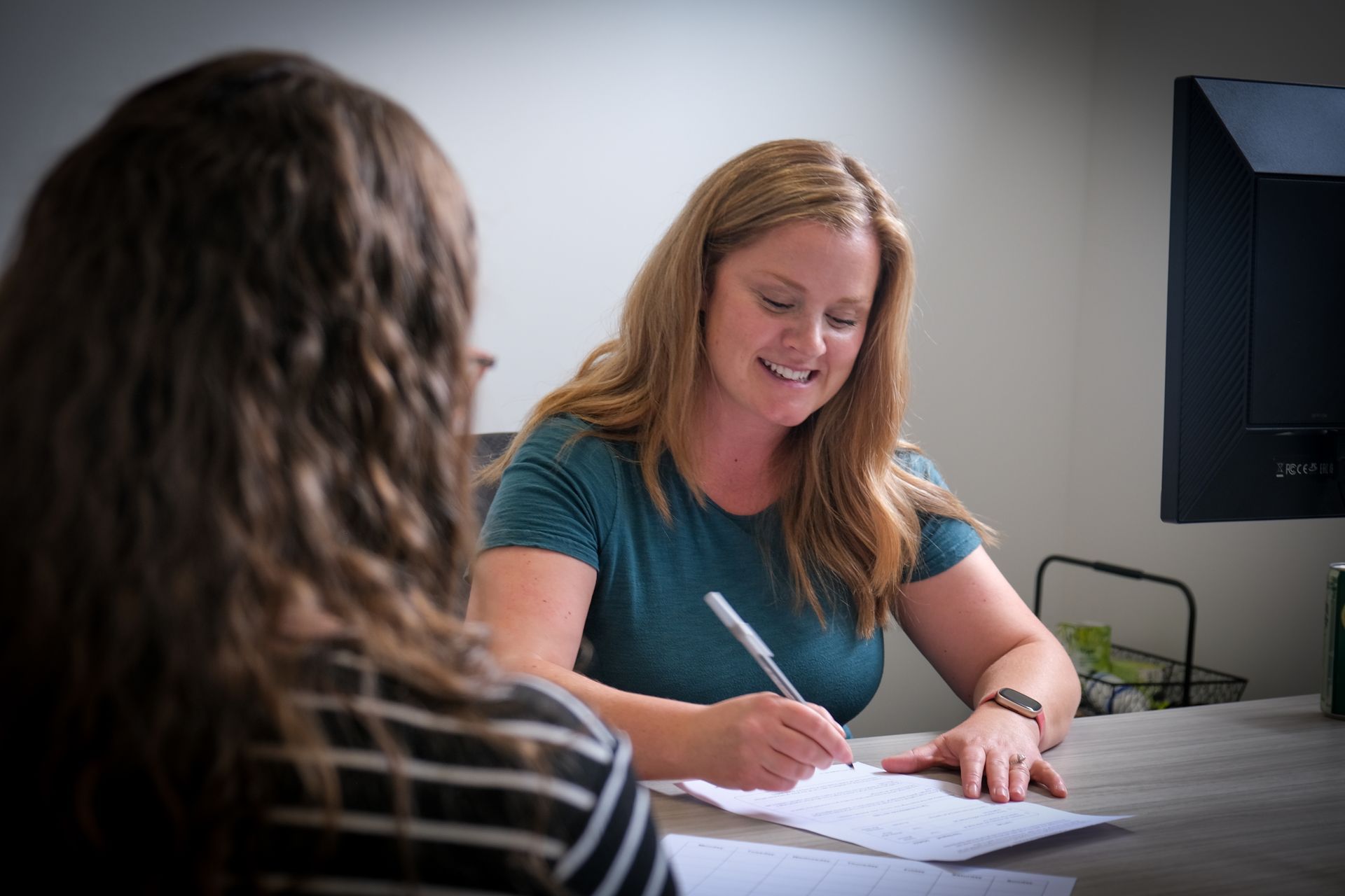 A person sits at a desk writing on a document while speaking with another person in an office setting.