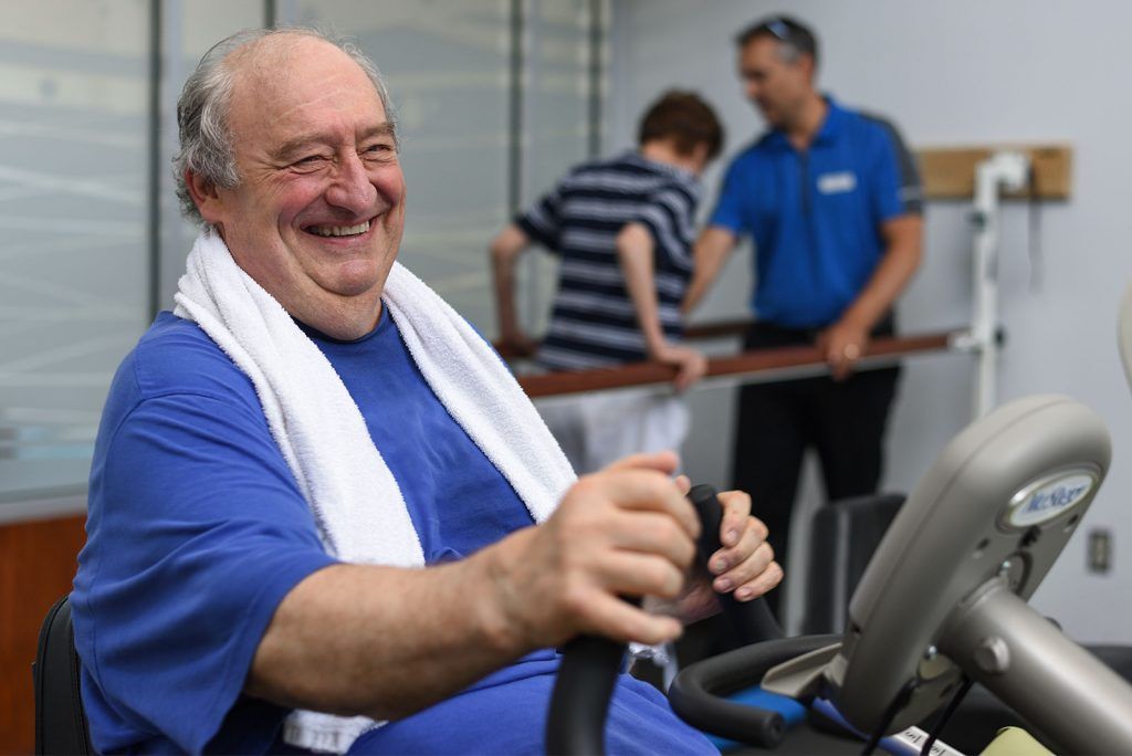 A person smiling while using a stationary exercise bike in a gym, with another person exercising in the background.