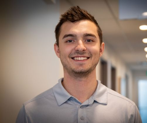 A person with short, dark hair and a light blue polo shirt smiling at the camera in a brightly lit hallway.
