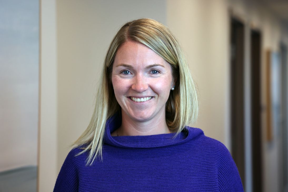 A person with blonde hair smiling while wearing a purple cowl-neck sweater in an office setting.