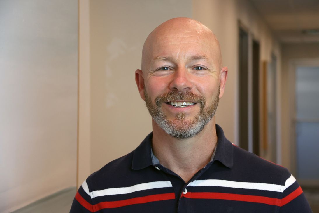 A smiling person with a shaved head and a beard, wearing a navy, red, and white striped polo shirt in an office setting.