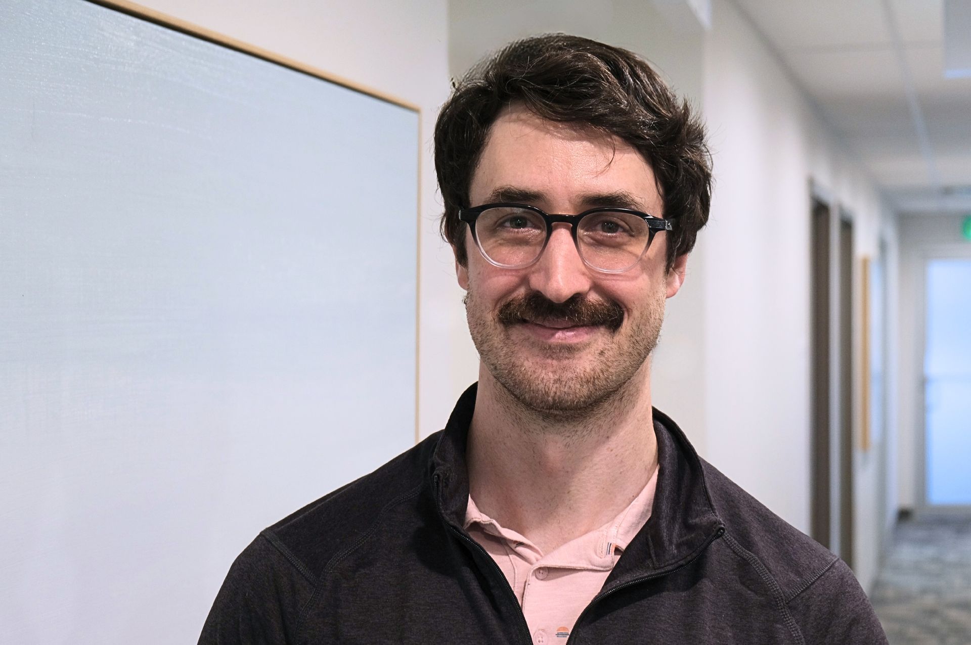 A person with wavy dark hair, glasses, and a mustache smiles while wearing a dark zip-up jacket in an office hallway.