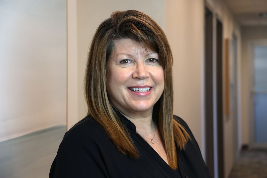 A person with shoulder-length brown hair wearing a black top, smiling in an office hallway.