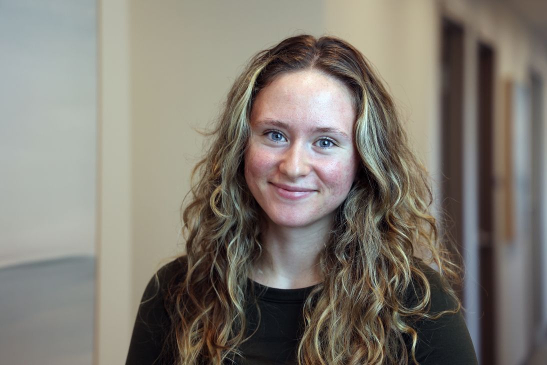 A person with long, wavy brown hair and blue eyes smiling at the camera in a brightly lit indoor space.