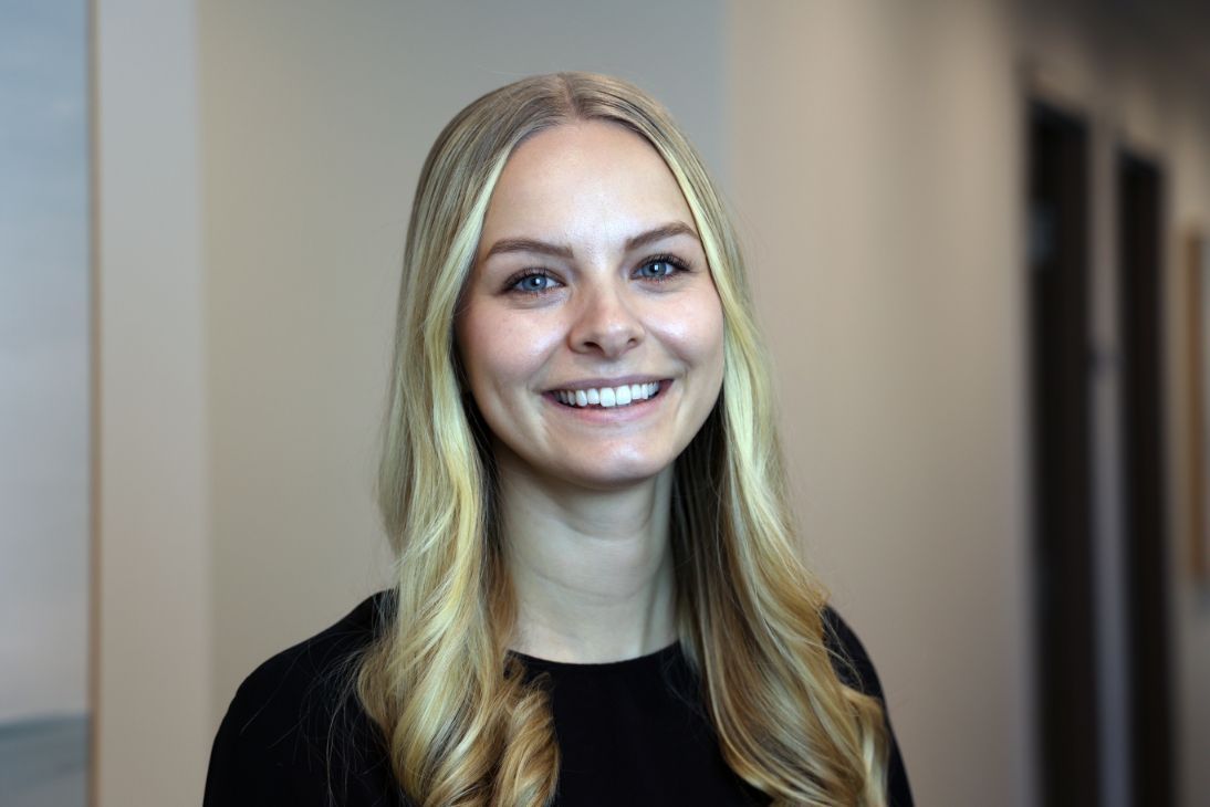 A smiling person with long blonde hair and a black top posing in front of a plain wall.