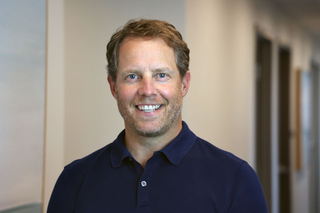A person with short wavy hair and a beard, wearing a navy polo shirt, smiling at the camera.