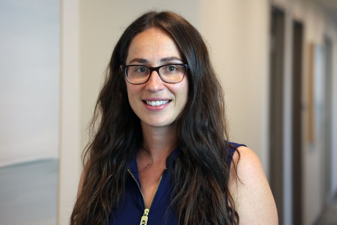 A smiling person with long, dark hair wearing glasses and a blue collared shirt stands in an office hallway.