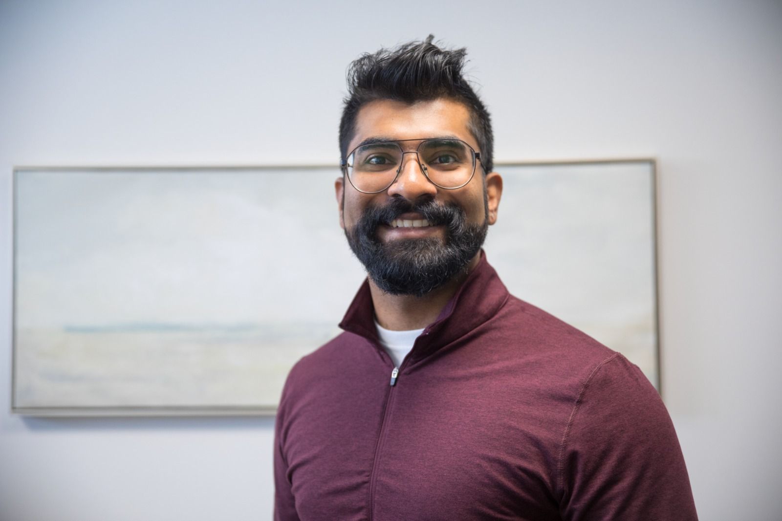 A person with a beard and glasses wearing a burgundy quarter-zip sweater smiles in front of a framed landscape painting.
