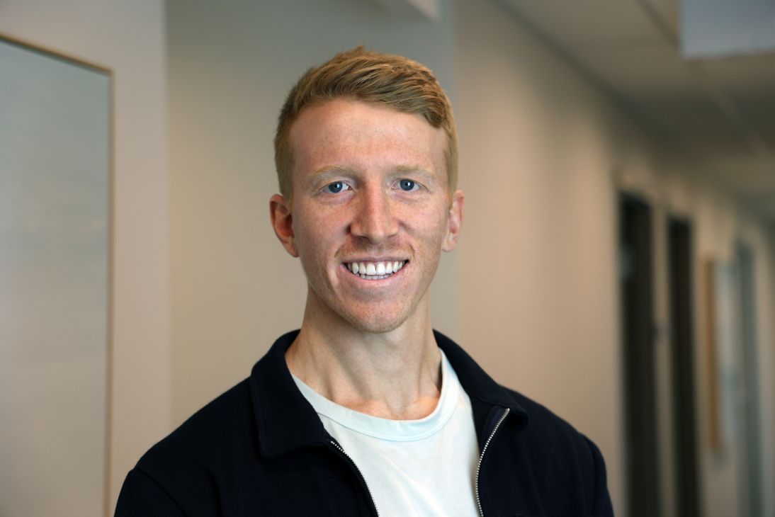 A smiling individual with light brown hair wears a white t-shirt and dark jacket in a hallway.