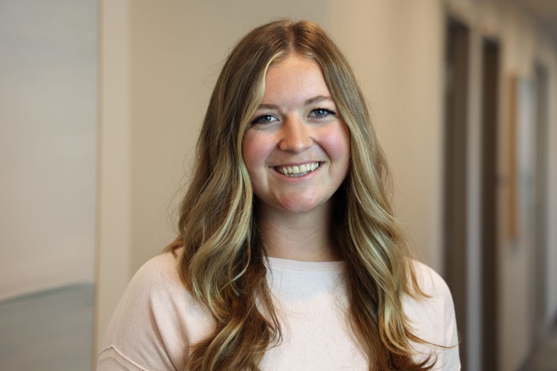 A person with long blonde hair and a friendly smile, wearing a light pink shirt, indoors against a neutral wall.
