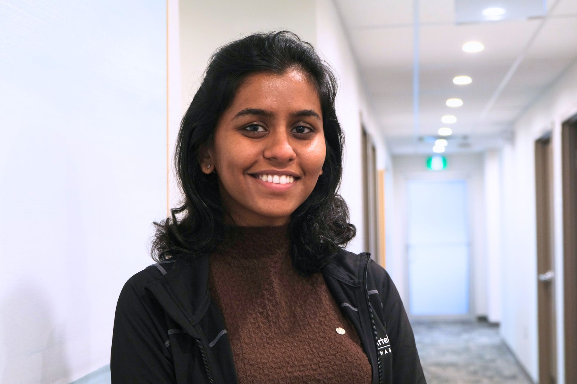 A person wearing a brown mock-neck top and black zip-up jacket smiles in a brightly lit hallway.