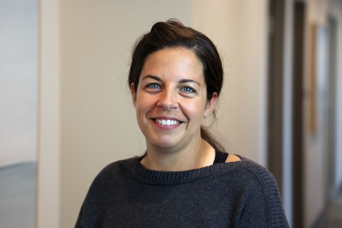 A person with dark hair pulled back smiles warmly while wearing a dark gray sweater in a brightly lit indoor space.