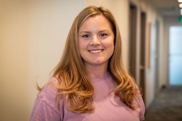 A person with shoulder-length blonde hair smiles while wearing a light purple shirt in an office hallway.