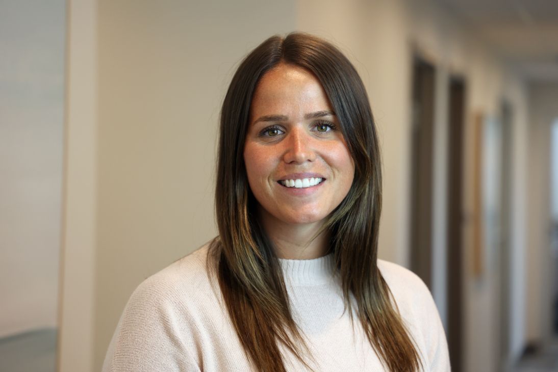 A person with long, dark hair wearing a light-colored sweater, smiling in a brightly lit office setting.
