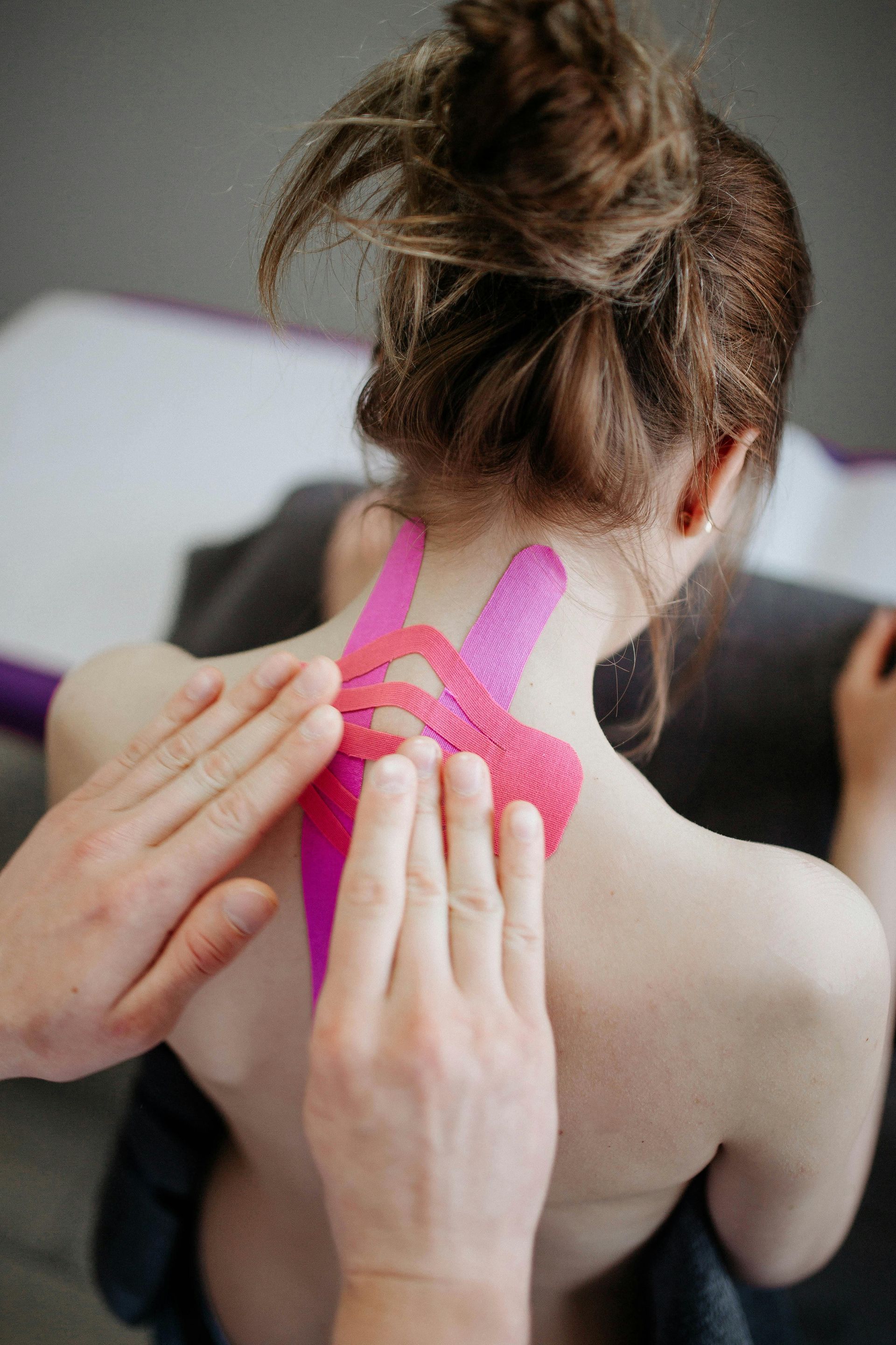 A therapist applying pink kinesiology tape to a person's upper back and neck area.