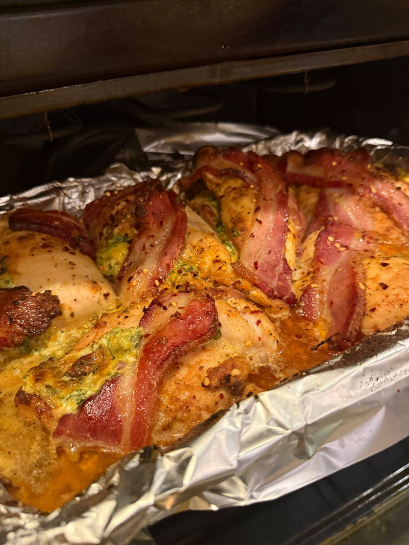 Chicken breasts wrapped in bacon, baking in an oven on a foil-lined pan.
