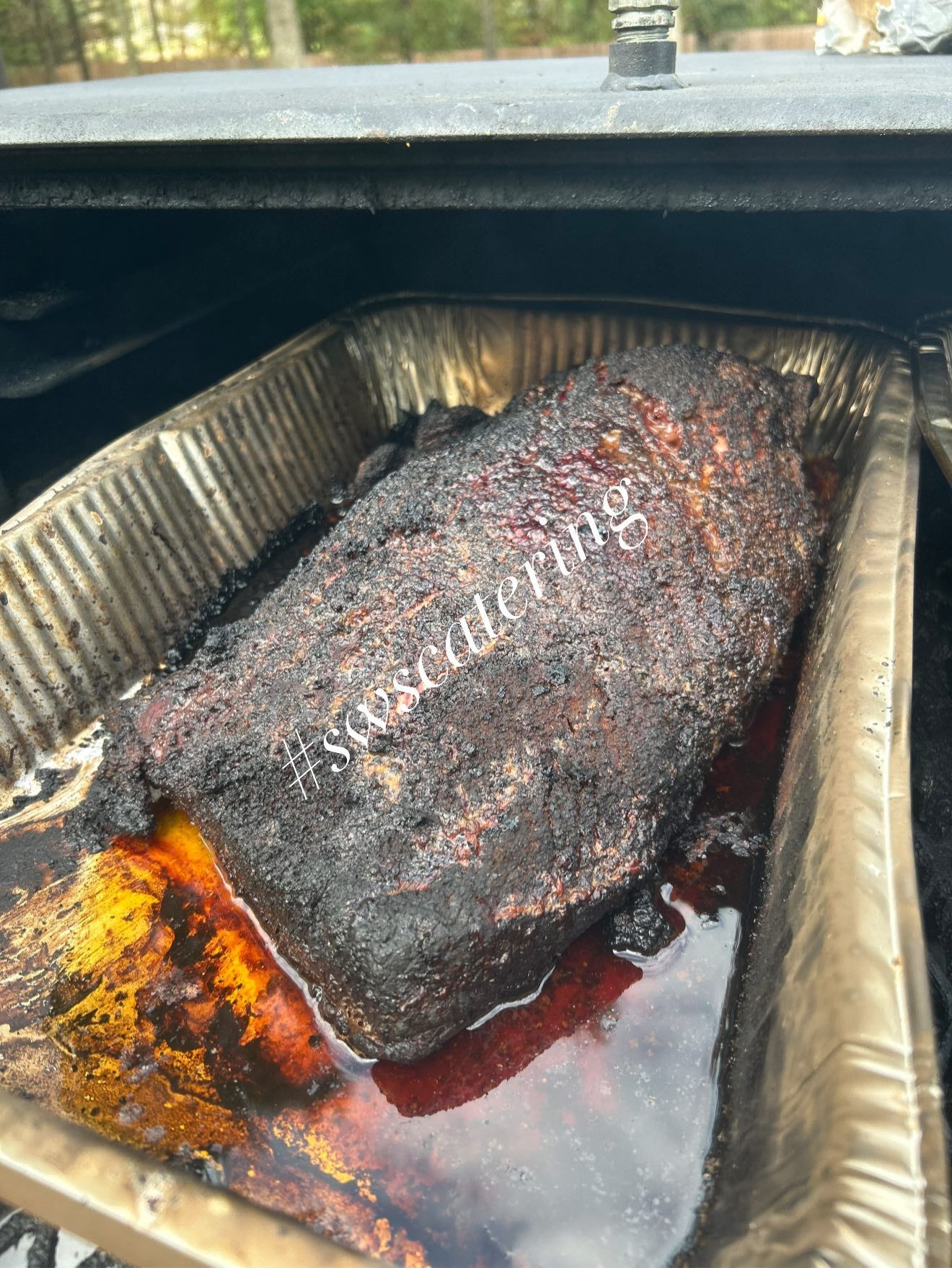 Brisket in a foil pan inside a smoker, with a dark bark and rendered fat.