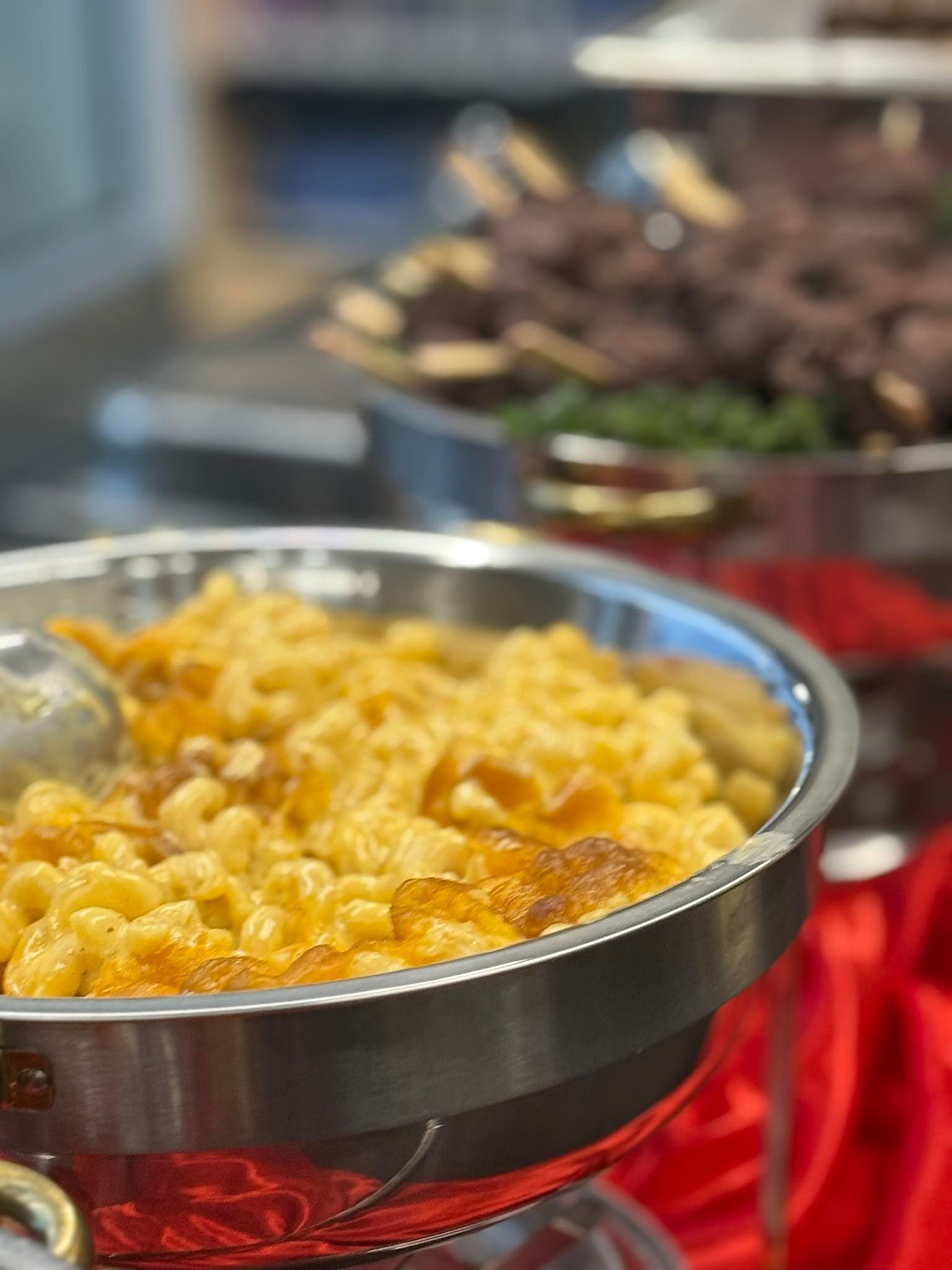 Macaroni and cheese in a silver chafing dish, with dark meat skewers in background.