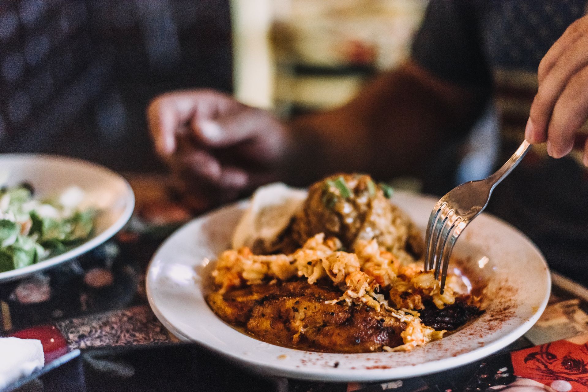 Person eating a meal on a white plate with rice, sauce, and meat; salad in a bowl on the side.