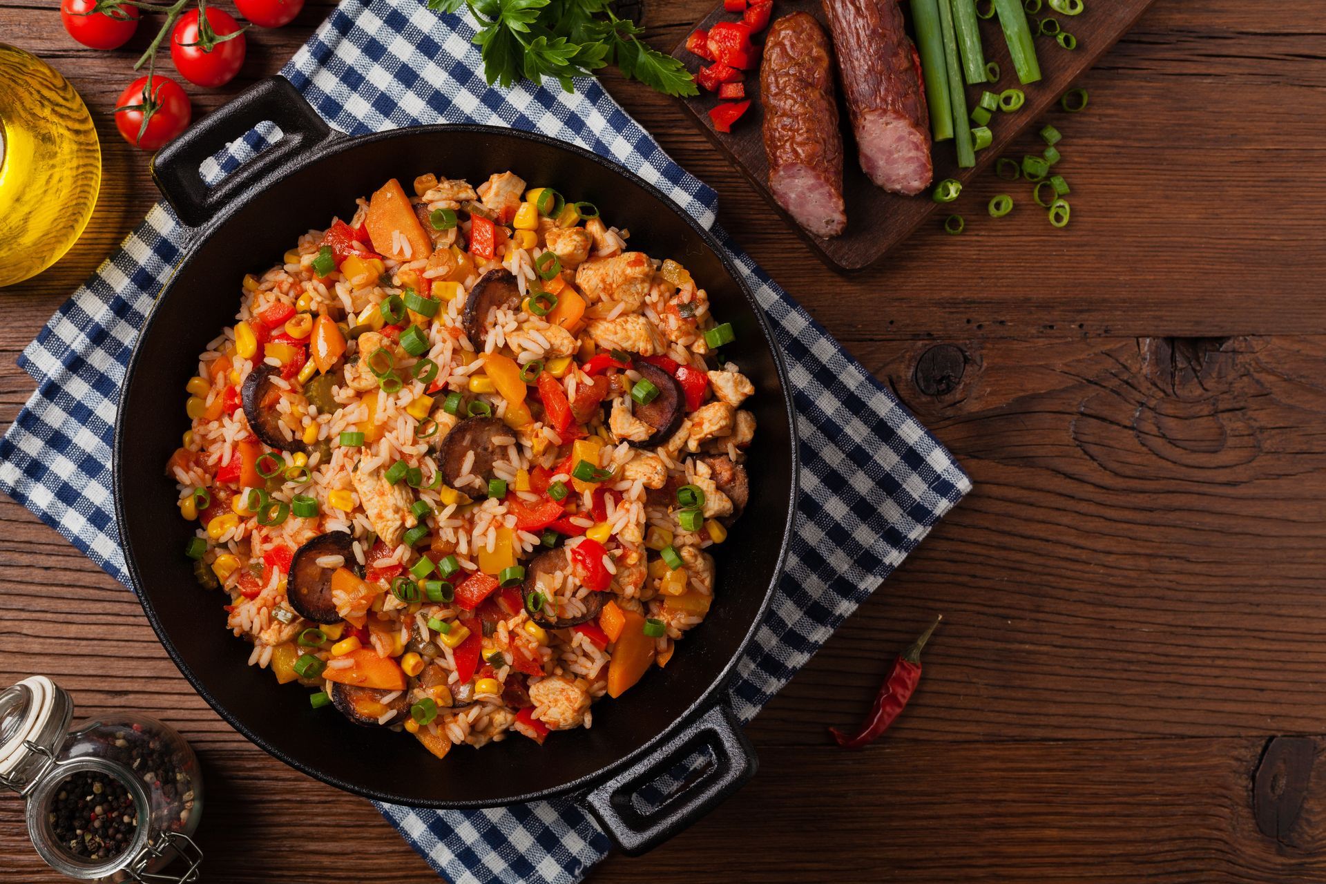 Chicken and rice dish with vegetables in a black pan on a wooden table, with sausage and garnishes.