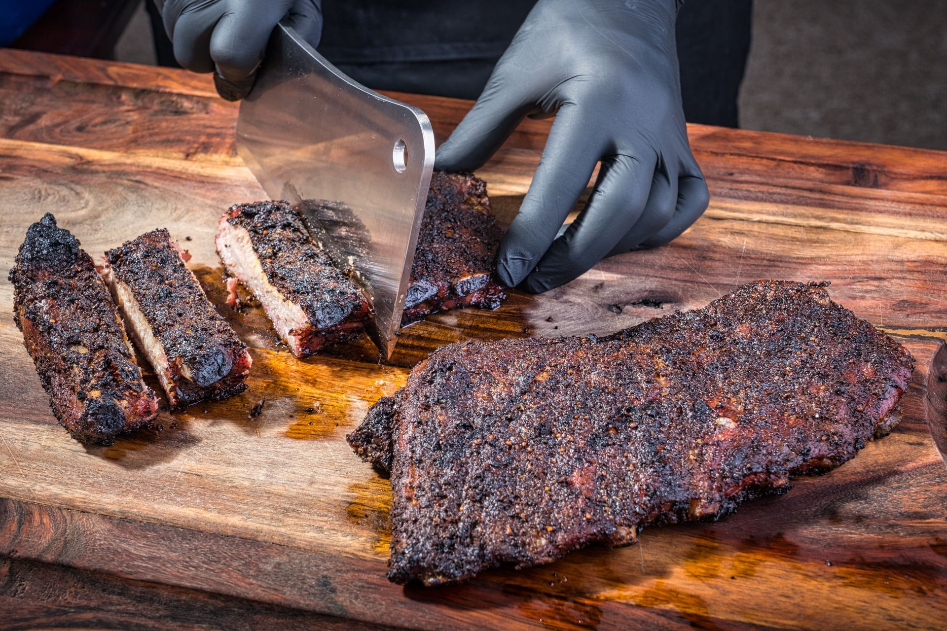 Person in black gloves slicing smoked ribs on a wooden cutting board with a cleaver.