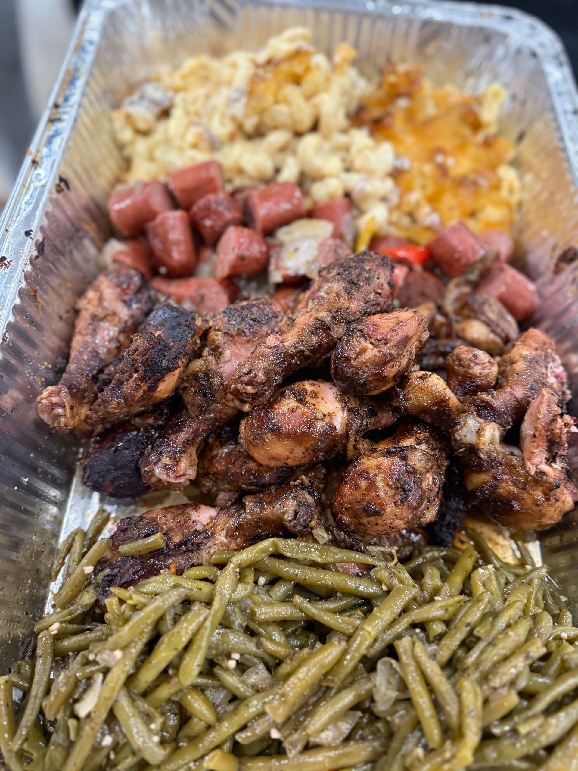 Aluminum tray of food with chicken drumsticks, green beans, mac and cheese, and sausage.