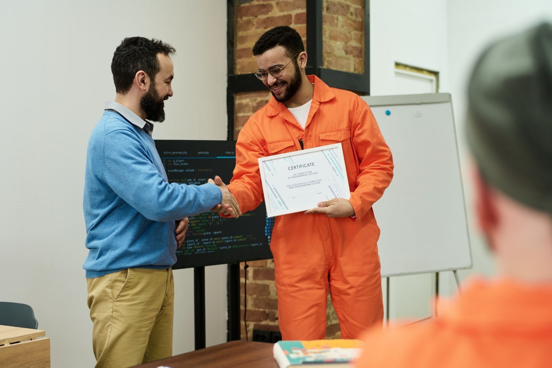 Two colleagues shaking hands, one in blue and one in orange, in an office setting with a whiteboard.